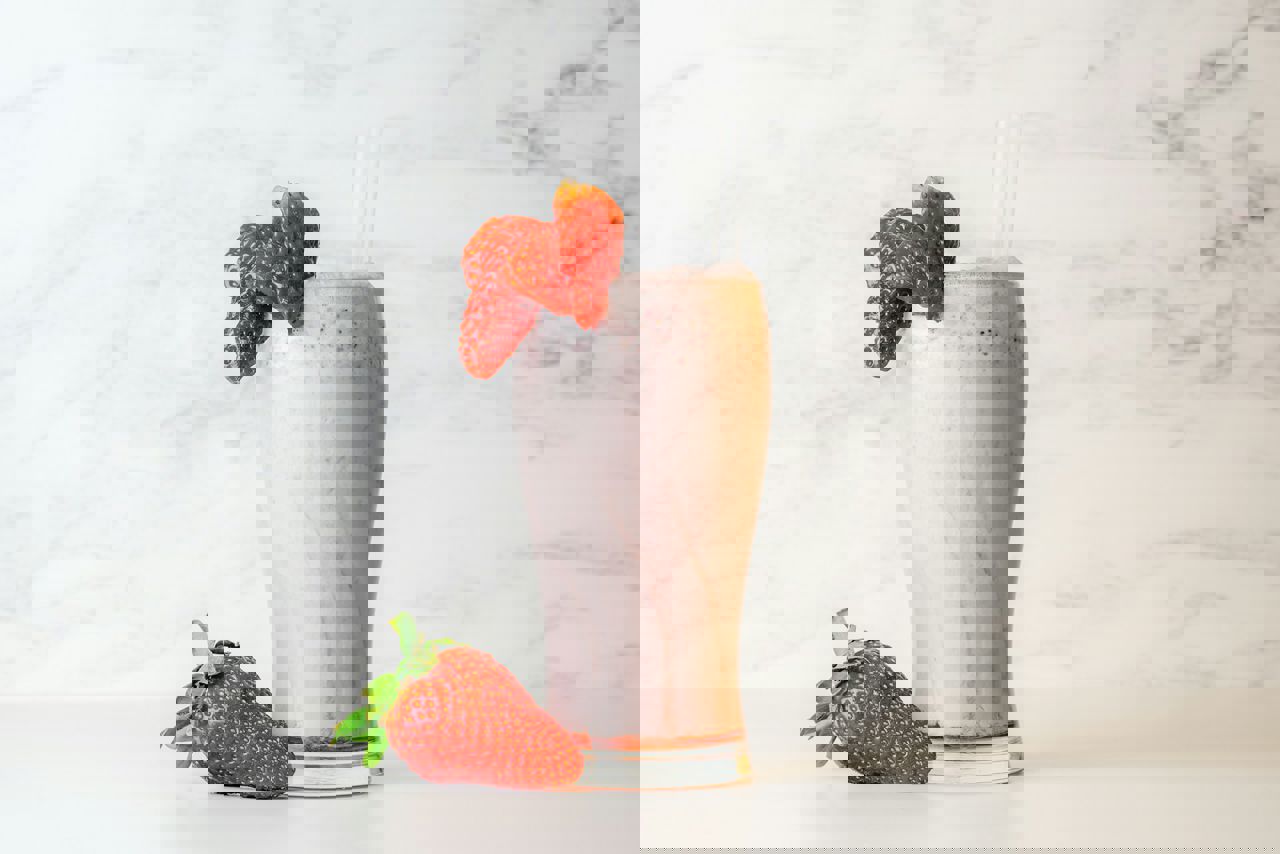 Et højt glas med jordbærmilkshake toppet med to sugerør og pyntet med friske jordbær, placeret på en lys baggrund.