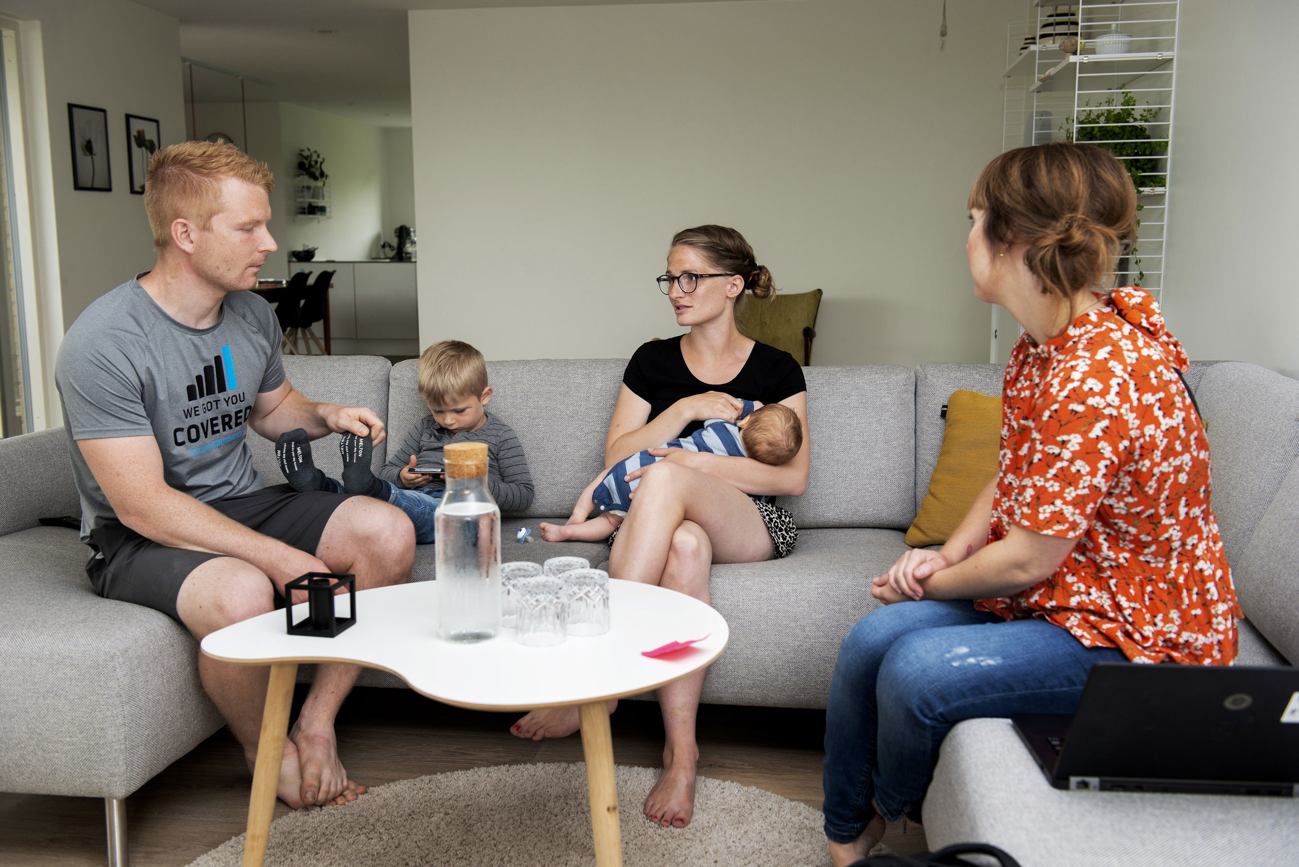 Mand, mindre dreng og kvinde med baby i sofa omkring sofabord - til højre sidder kvinde i blomstret bluse