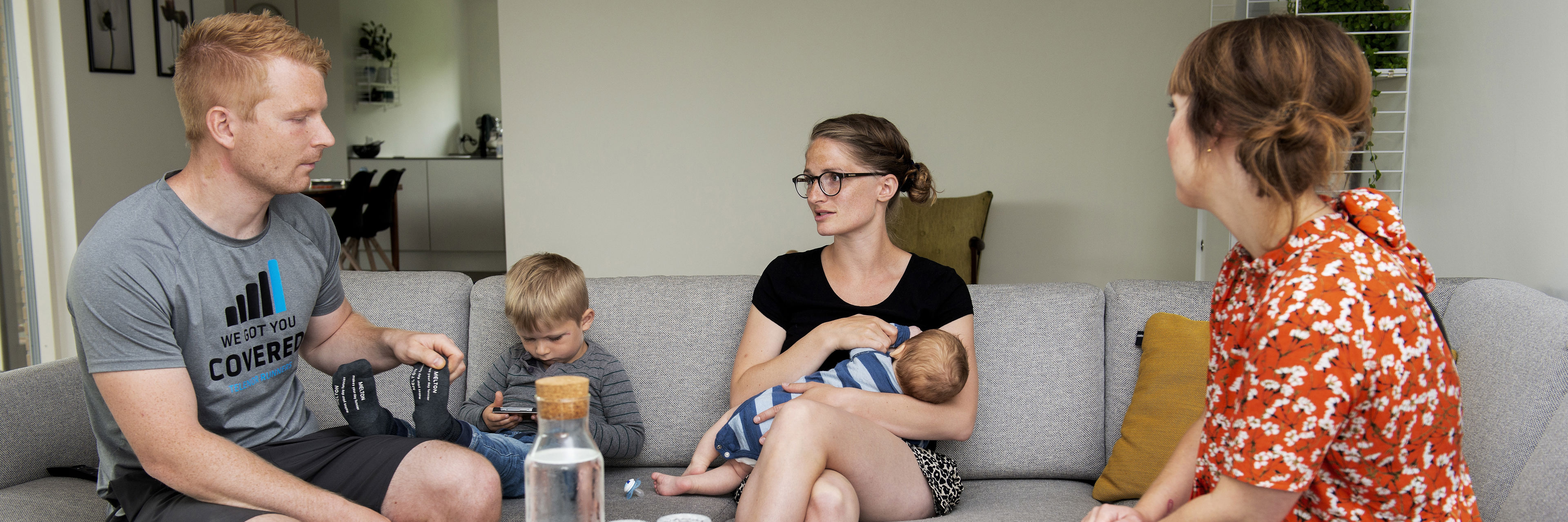 Mand, mindre dreng og kvinde med baby i sofa omkring sofabord - til højre sidder kvinde i blomstret bluse