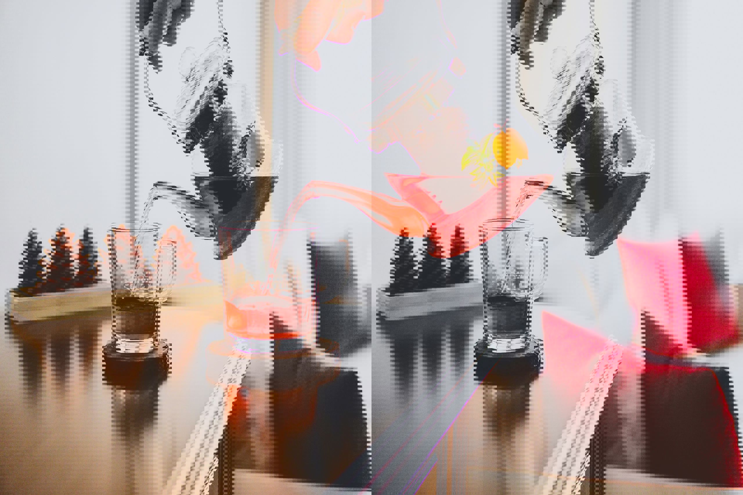 En person hælder rød frugtte fra en glaskande ned i en glaskop på et bord, med pyntekogler og puder i baggrunden.