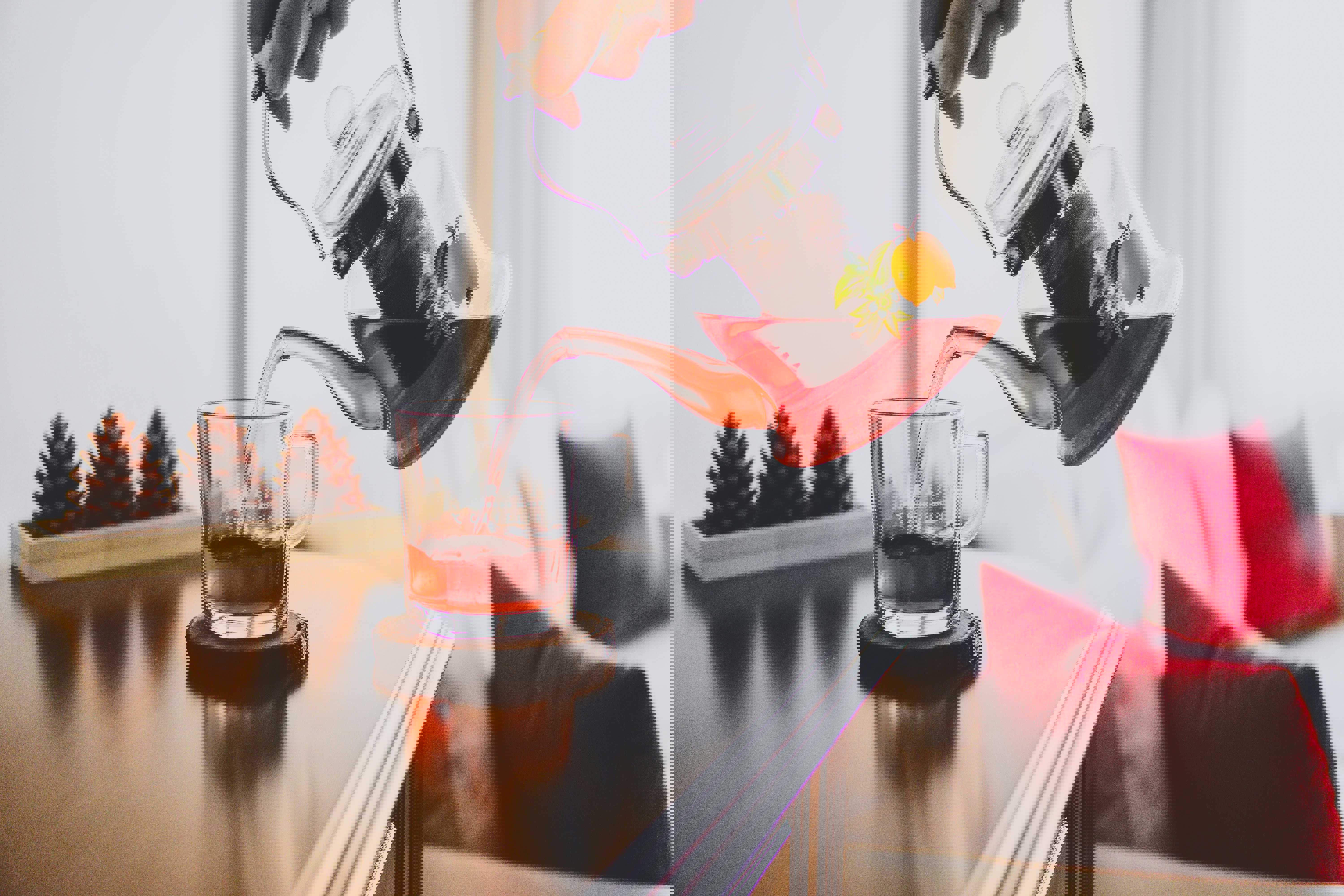 En person hælder rød frugtte fra en glaskande ned i en glaskop på et bord, med pyntekogler og puder i baggrunden.