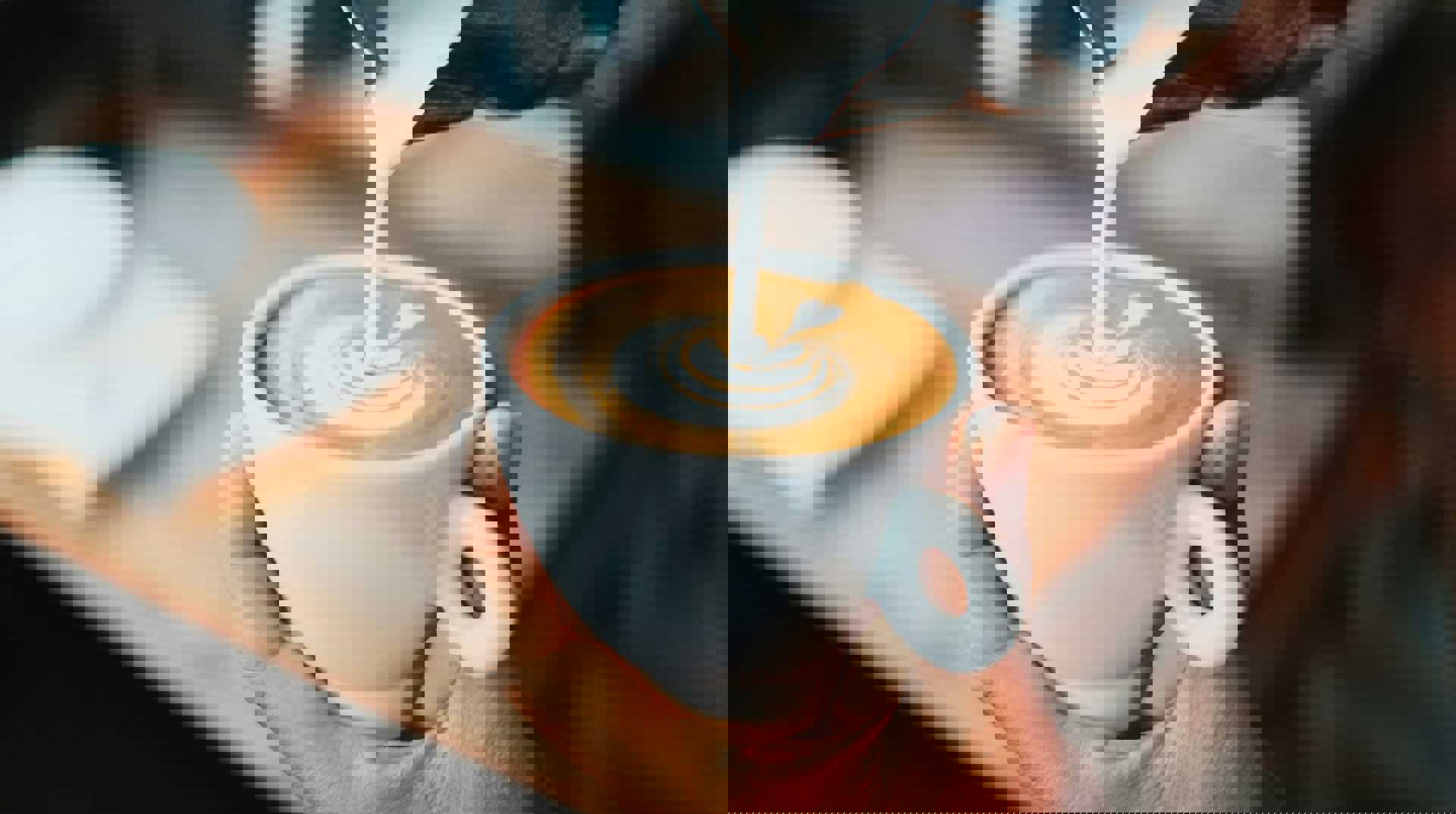 Nærbillede af en hånd, der holder en hvid kop cappuccino, mens mælk hældes i og danner latte art.