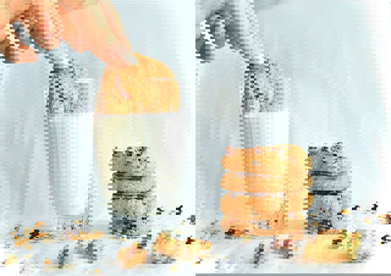 En hånd dypper en småkage i et glas mælk, mens en stak af fire småkager ligger ved siden af på et hvidt underlag med krummer omkring.